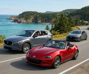 Obraz przedstawia samochody marki Mazda w realistycznych, naturalnych warunkach drogowych